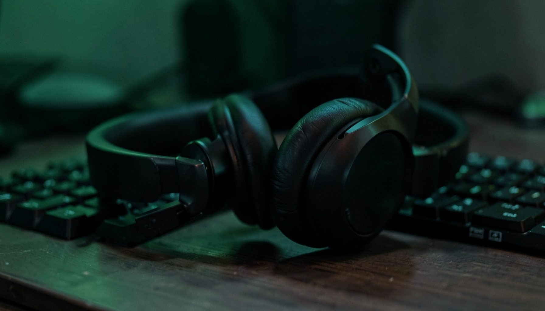 Dark over-ear headphones resting on a desk beside a keyboard, moody ambient lighting with subtle green glow