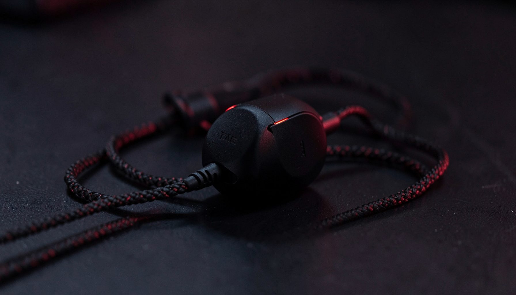 Compact in-ear gaming headset with braided cable on a dark surface, subtle red accent lighting